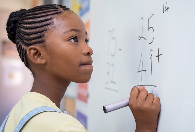 Girl with braided hair thoughtfully solves a math problem on a whiteboard, focusing on subtraction and addition equations in a classroom setting.