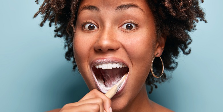 Smiling woman with curly hair brushes her teeth, showcasing oral hygiene. The blue background emphasizes her enthusiasm for dental care.